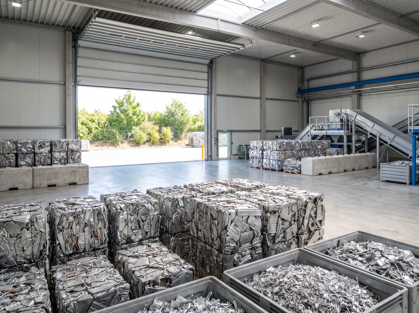 Baled aluminium scrap stacked inside a bright recovery hall, with the roller door open to daylight.